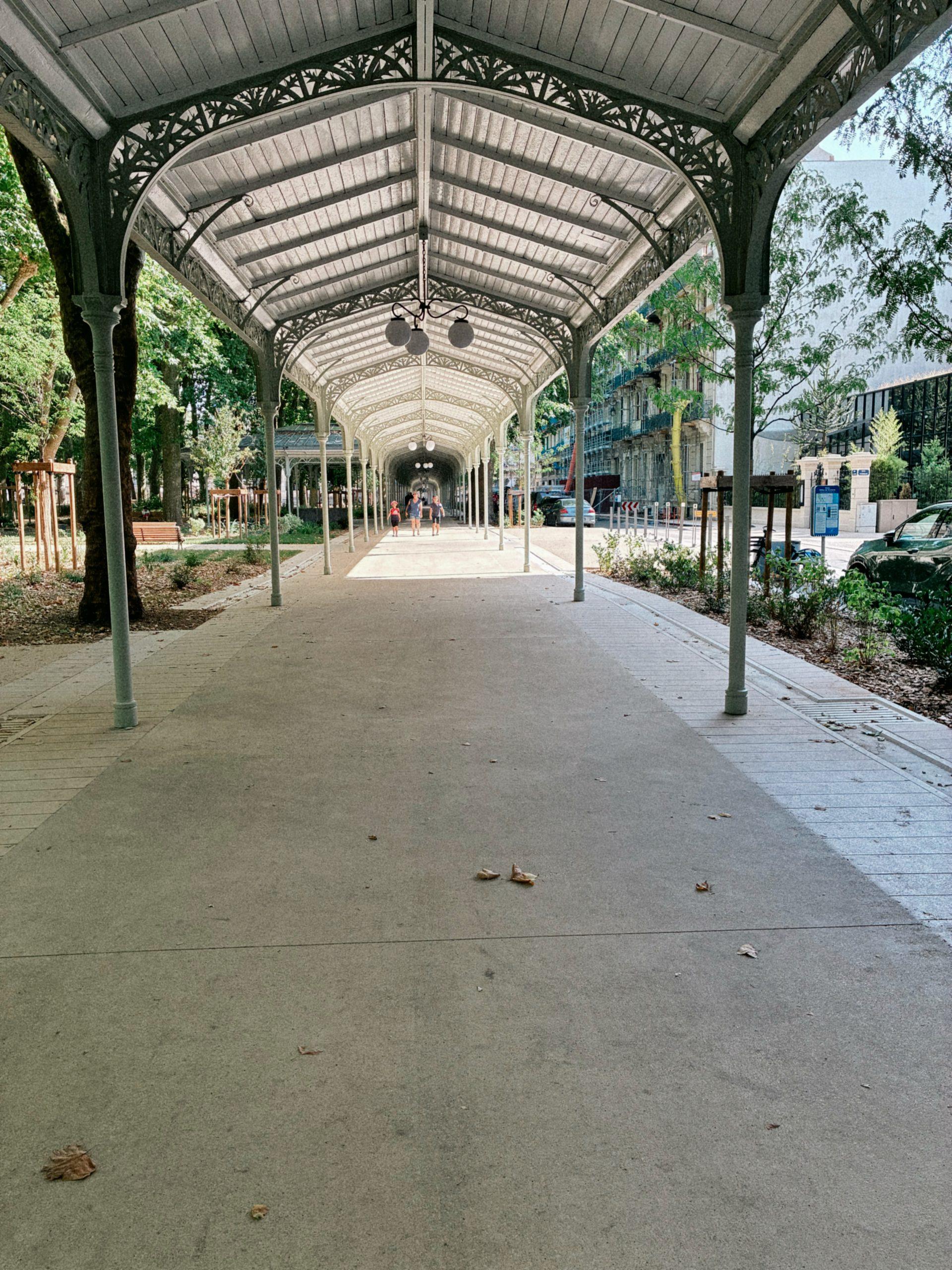 Longue galerie couverte en fer forgé ornementé menant à une allée bordée d'arbres, avec des promeneurs au loin