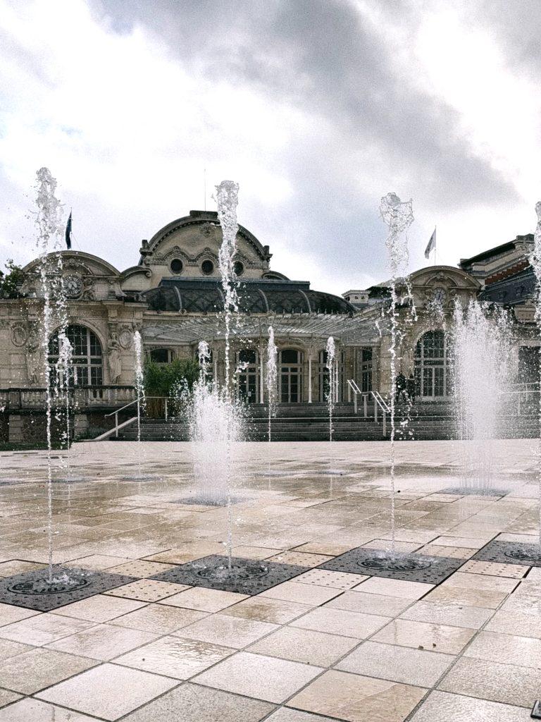 Jets d'eau jaillissant d'une place pavée devant un bâtiment ancien à coupole et verrière