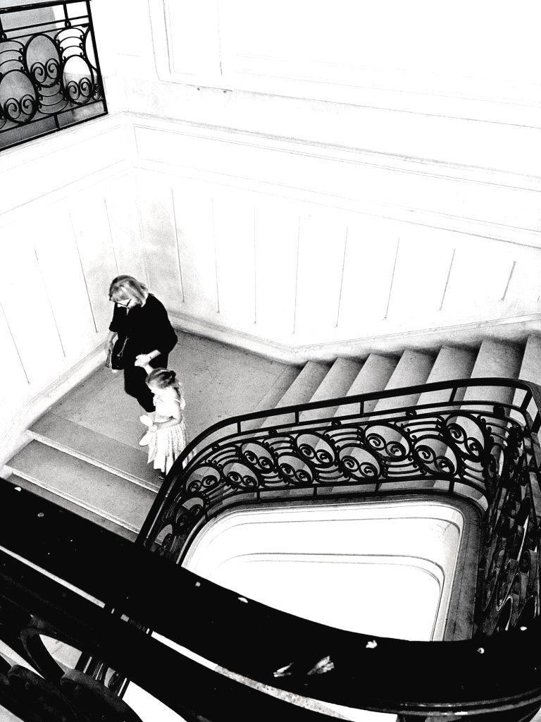 Photo noir et blanc façon Cartier-Bresson : escalier à rampe en fer forgé vu en plongée, une femme et une fillette descendent