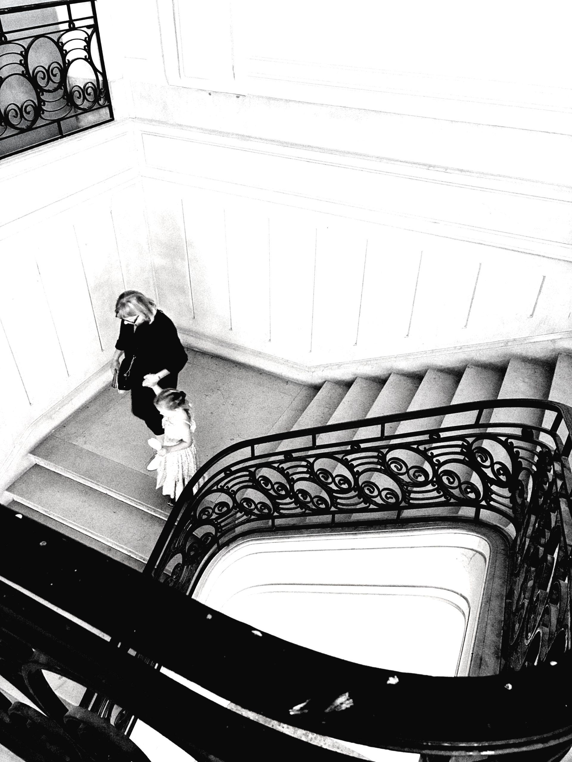 Photo noir et blanc façon Cartier-Bresson : escalier à rampe en fer forgé vu en plongée, une femme et une fillette descendent